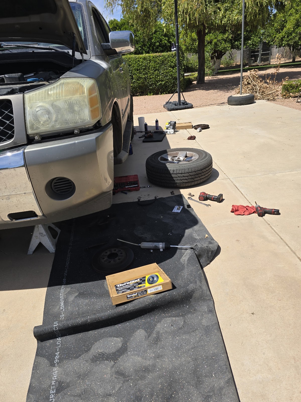 Front brake job in a sunny driveway: a silver pickup on jack stands with the hood up and front wheel off, the wheel resting on the concrete, an old rotor and grease gun on a black rubber mat, a new Duralast brake rotor still in its box, red cordless power tools and a socket set nearby